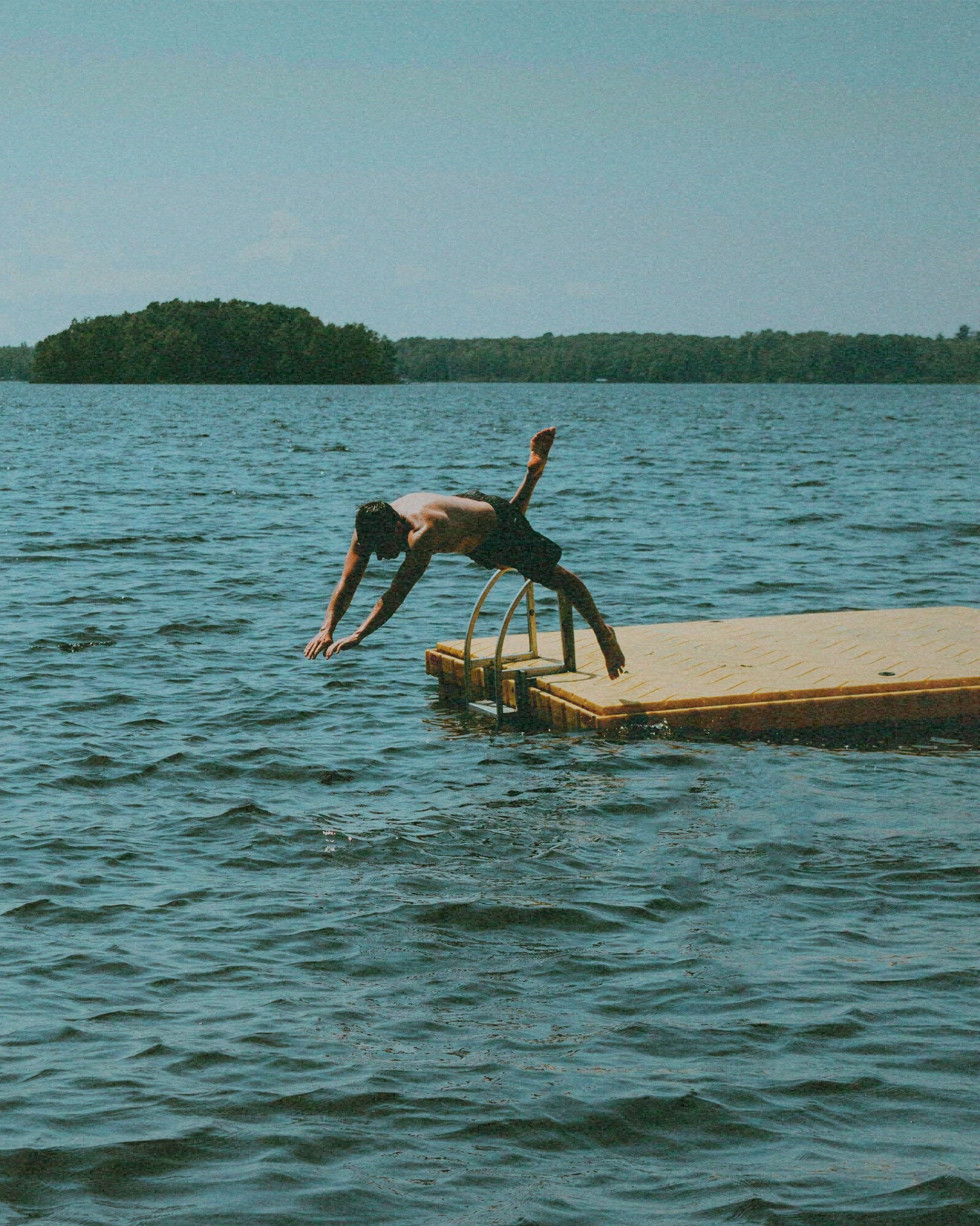 A man diving off a floating dock into a lake, a forested island beyond